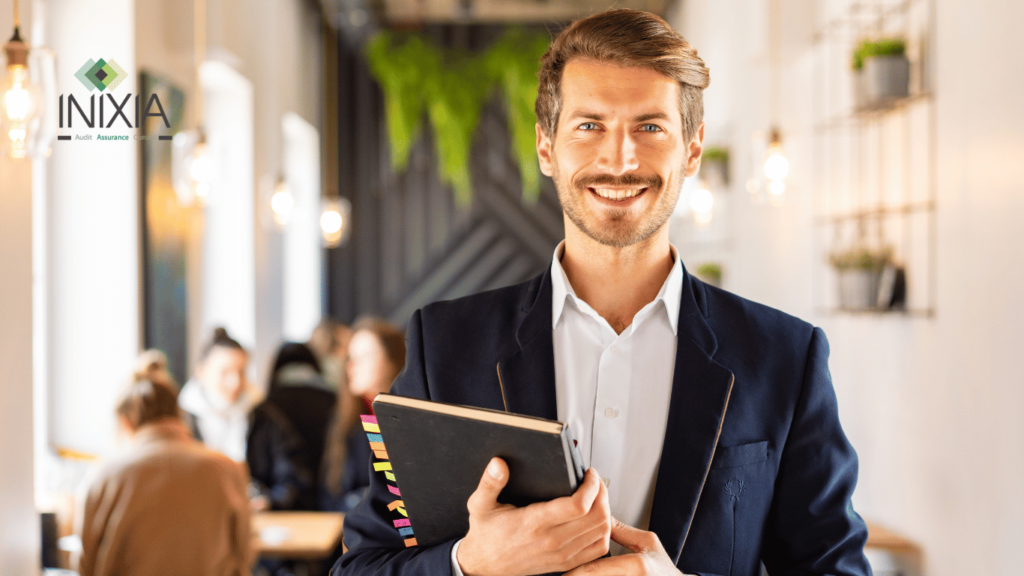 Un homme d'affaires souriant, tenant un agenda ou un cahier, se tient dans un espace de café moderne avec des plantes suspendues et des étagères décoratives. Le logo d'INIXIA est affiché en haut à gauche, indiquant une affiliation professionnelle avec la société d'audit, d'assurance et de conseil.