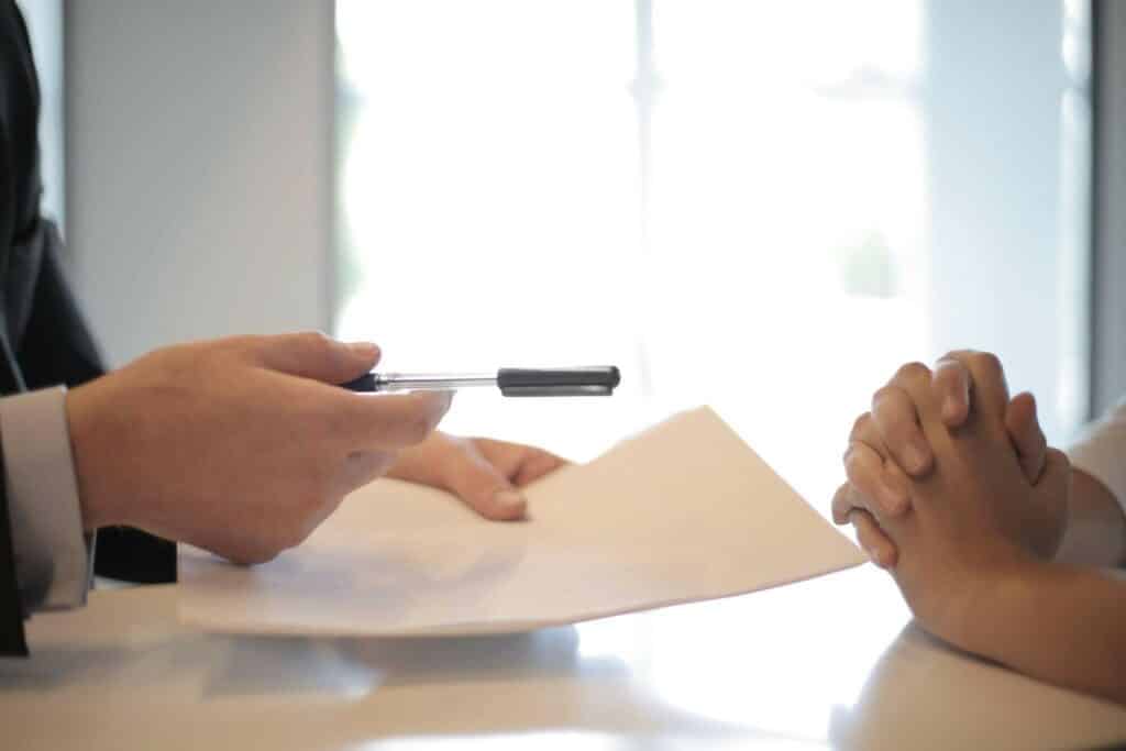Un professionnel tend un stylo et un document à une autre personne qui attend avec les mains jointes, prête à signer.